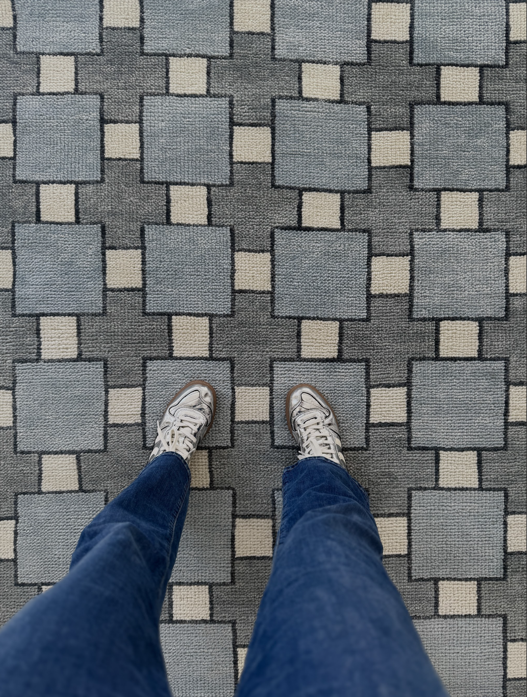 Person wearing blue jeans and white sneakers standing on a patterned gray and beige carpet.