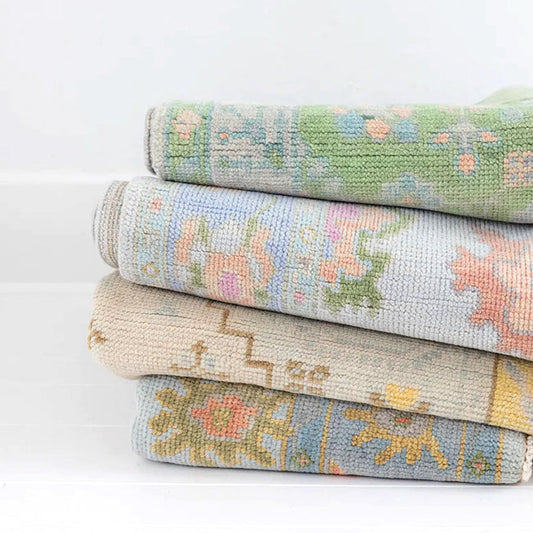 Stack of four rolled handcrafted area rugs featuring soft pastel floral, Oushak-inspired, and geometric patterns in shades of sage green, blue, ivory, blush, and gold.