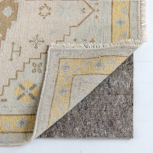 Corner of a handcrafted Oushak-style wool rug folded back to reveal a premium felt rug pad underneath, showcasing the rug's woven backing and non-slip cushioning on a hardwood floor.