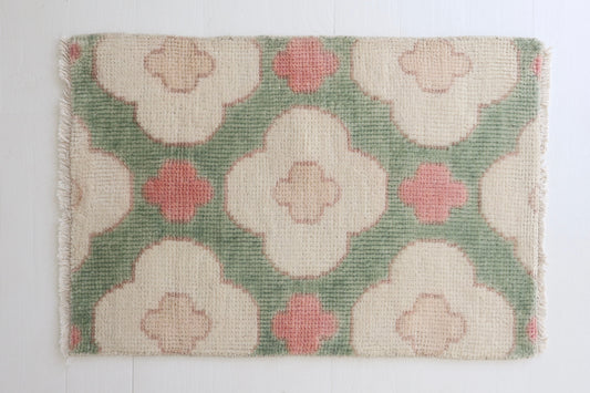 A rectangular woven rug with a muted green background and large cream floral motifs, accented by dusty pink quatrefoil shapes. The rug has fringed edges and is photographed flat against a white background.
