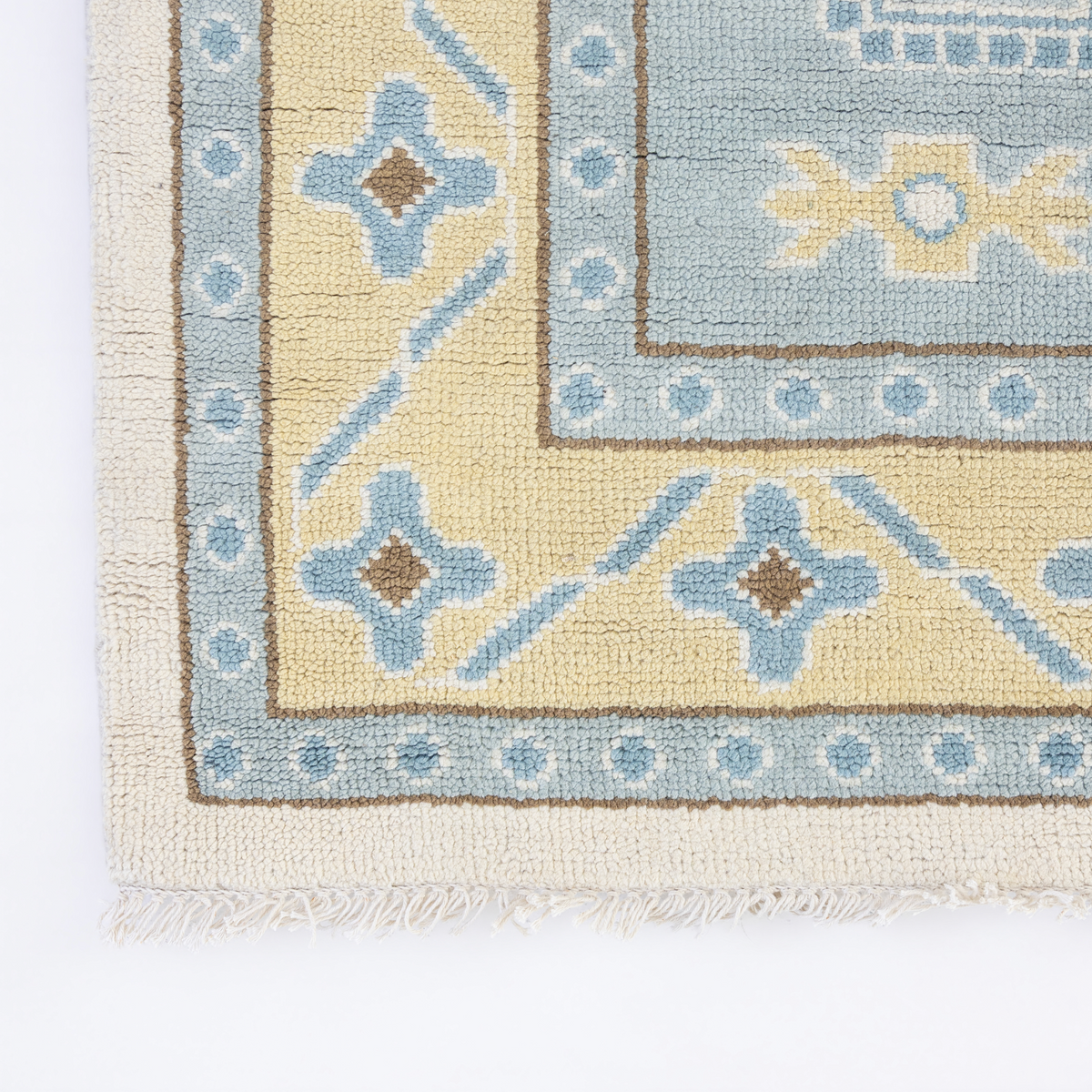 Close-up of a patterned rug corner showing cream fringe, a beige field, a light blue border with brown and blue geometric motifs, and a pale blue inner section on a white background.