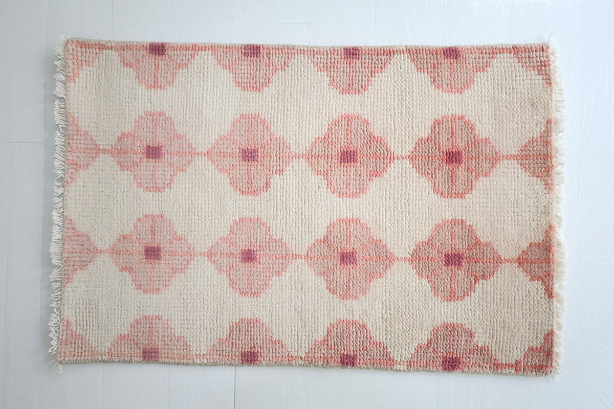 A rectangular woven rug with a cream background and a repeating dusty pink geometric diamond pattern, finished with short fringe on the left and right edges, laid flat on a white tiled floor.