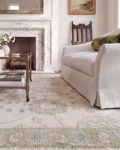Essie hand-knotted floral area rug with an ivory background and muted blue and green accents beside a cream sofa.