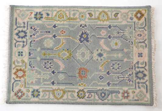 A square handwoven rug with a muted blue-gray field, cream border, and multicolored floral and geometric motifs in blue, yellow, orange, and pink, finished with white fringe on the sides.