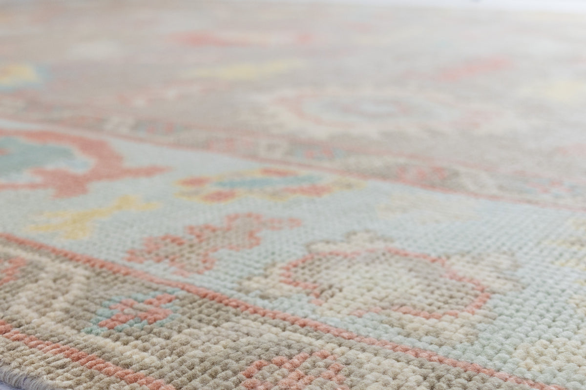 A close-up, angled view of a textured area rug with a traditional floral pattern in soft pastel tones of pink, cream, light blue, and yellow on a warm beige background.
