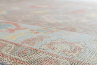 A close-up, angled view of a textured area rug with a traditional floral pattern in soft pastel tones of pink, cream, light blue, and yellow on a warm beige background.
