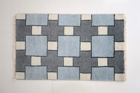A rectangular woven rug with a geometric block pattern in light blue, dark gray, and off-white, featuring short fringe on the sides, displayed against a white background.