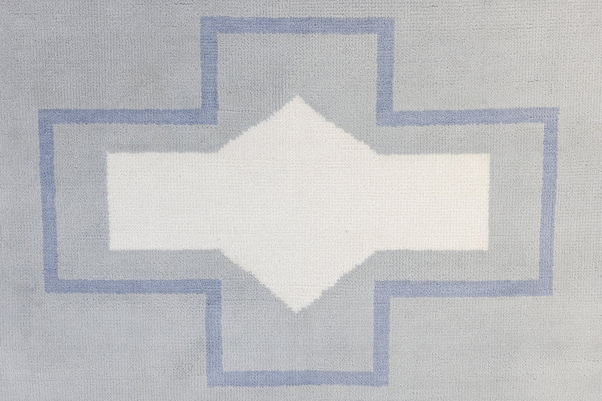 A close-up of a woven textile with a geometric pattern featuring a white diamond in the center, surrounded by a light gray cross shape outlined in dusty blue.