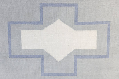 A close-up of a woven textile with a geometric pattern featuring a white diamond in the center, surrounded by a light gray cross shape outlined in dusty blue.