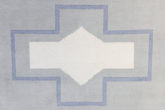 A close-up of a woven textile with a geometric pattern featuring a white diamond in the center, surrounded by a light gray cross shape outlined in dusty blue.