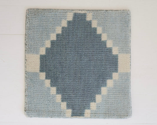 A square woven rug with a geometric diamond pattern in slate blue and cream, displayed flat against a white paneled wall.