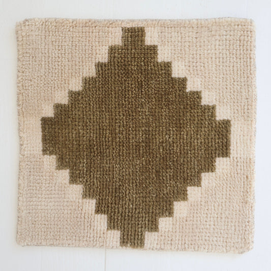 A square woven rug with a geometric diamond pattern in dark brown on a cream background, displayed against a white wall.